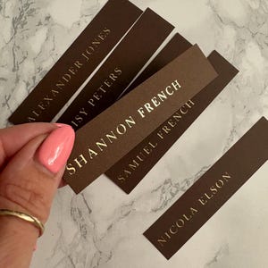 以下が含まれることがあります： 金色の文字が入った茶色の長方形の名札。 「Shannon French」や「Alexander Jones」などの名前が表示されています。 ピンク色のマニキュアを塗った爪と金の指輪をした手で名札を持っています。 背景は白とグレーの大理石模様です。