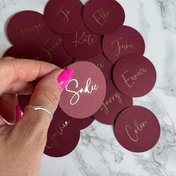 May include: Burgundy-colored circular place cards with gold and white calligraphy names. One card is held by a hand with pink nail polish. The names include George, Elle, Kate, June, Eunice, Garry, Colin, and Sadie.