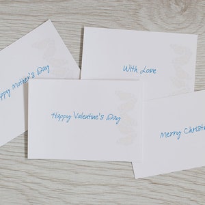 May include: Four white cards with blue handwritten text. The cards have a decorative design in the corner. The text on the cards reads "Happy Mother's Day", "Happy Valentine's Day", "With Love", and "Merry Christmas".