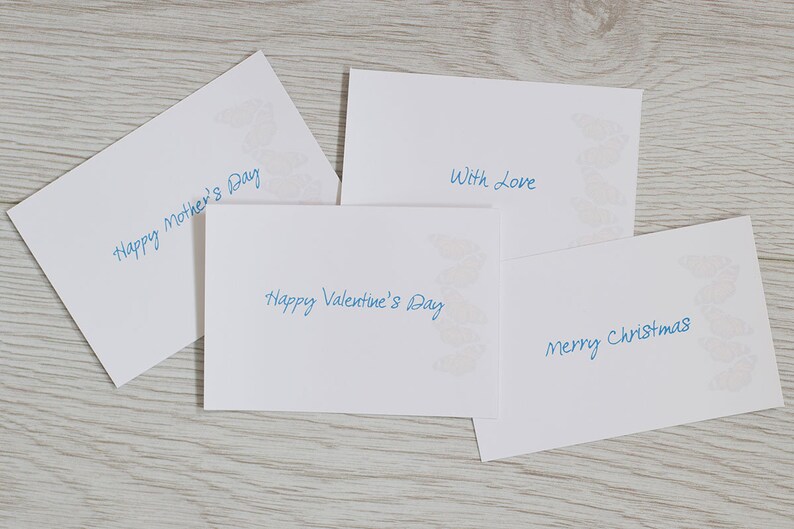 May include: Four white cards with blue handwritten text. The cards have a decorative design in the corner. The text on the cards reads "Happy Mother's Day", "Happy Valentine's Day", "With Love", and "Merry Christmas".
