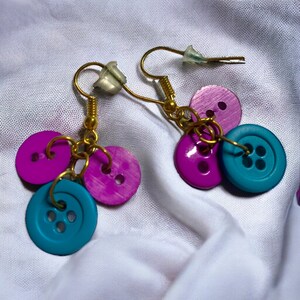 May include: A pair of earrings made with purple and teal buttons. The earrings are made with gold-coloured wire and have a simple design.