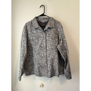 Może przedstawiać: Szara koszula zapinana na guziki z wzorem róż. Koszula z długim rękawem wisi na czarnym wieszaku na białej ścianie. Koszula ma kołnierzyk i dwie przednie kieszenie.