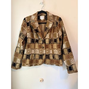May include: A vintage blazer with a geometric pattern in shades of brown, tan, and black. The jacket has a notched collar, two buttons, and long sleeves. The blazer is hanging on a black hanger against a white wall.