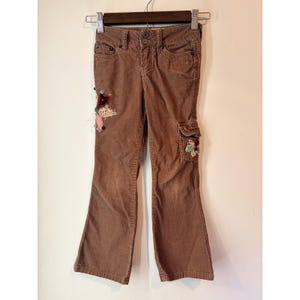 May include: Brown corduroy pants with a flared leg. The pants have a button and zipper closure, front and back pockets, and a cargo pocket on the right leg. Decorative floral and butterfly appliques are sewn on the left leg and cargo pocket.