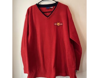 May include: A red long-sleeved V-neck sweatshirt with a navy blue trim around the neckline. The sweatshirt features a small embroidered patch with the text "POOH" in yellow and orange. The garment is hanging on a black hanger.