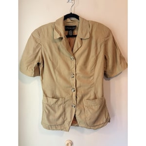 Puede incluir: Una blazer beige de manga corta con cuello con solapas, tres botones y dos bolsillos delanteros. La blazer está colgada de una percha negra contra una pared blanca. El nombre de la marca es visible en la etiqueta.