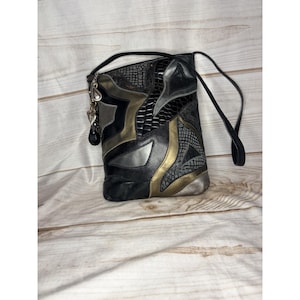 Könnte beinhalten: Schwarze, goldene und silberne Umhängetasche mit Patchwork-Design. Die Tasche hat einen langen schwarzen Riemen und einen dekorativen Anhänger. Die Tasche hat eine rechteckige Form und besteht aus verschiedenen Texturen.
