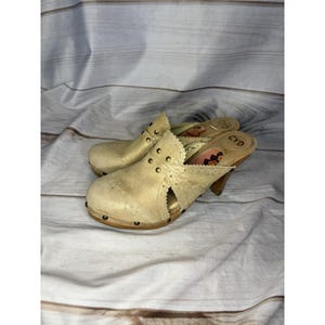 Könnte beinhalten: Ein Paar beigefarbene Wildleder-Clogs mit Holzsohlen und hohen Absätzen. Die Clogs haben dekorative Metallnieten und Ausschnittdetails. Die Innenseite der Clogs zeigt ein Blumenmuster und die Buchstaben "CYD".