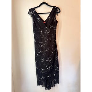 Puede incluir: Vestido negro sin mangas con cuello en V y estampado floral. El vestido tiene detalles de lazo en los hombros y un dobladillo asimétrico. El diseño floral presenta flores blancas y grises.
