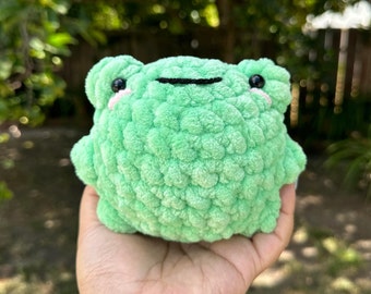 以下が含まれることがあります： 黒い目と黒い口をした緑色の毛糸で編まれたカエルのぬいぐるみ。カエルは人の手に乗っています。