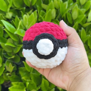 May include: A crocheted Poké Ball toy, featuring a red top, a black band, and a white lower section. The soft toy is held in a hand, with a background of green leaves. The ball is designed to resemble the iconic item.