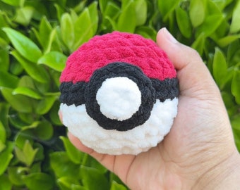 May include: A crocheted Poké Ball toy, featuring a red top, a black band, and a white lower section. The soft toy is held in a hand, with a background of green leaves. The ball is designed to resemble the iconic item.