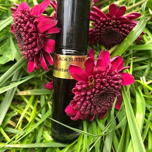 May include: A black glass bottle with a black cap, labeled "Black Butter", rests among vibrant red flowers and green grass. The bottle appears to contain a dark liquid. The overall aesthetic is natural and botanical.
