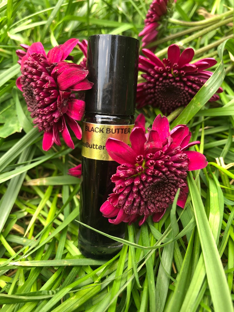 May include: A black glass bottle with a black cap, labeled "Black Butter", rests among vibrant red flowers and green grass. The bottle appears to contain a dark liquid. The overall aesthetic is natural and botanical.