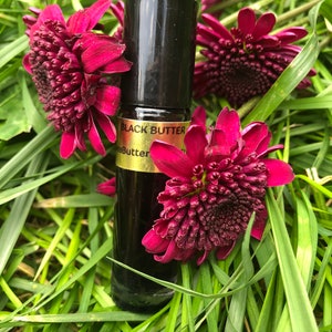 May include: A black glass bottle with a black cap, labeled "Black Butter", rests among vibrant red flowers and green grass. The bottle appears to contain a dark liquid. The overall aesthetic is natural and botanical.