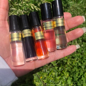 May include: Five small, clear glass roll-on bottles with black caps are held in a hand. The bottles contain various colored liquids, including pink, orange, and clear. Each bottle has a gold label with text, including "PUSS," "BLACK," "PINK SUGAR," and "BUTT NAKED."