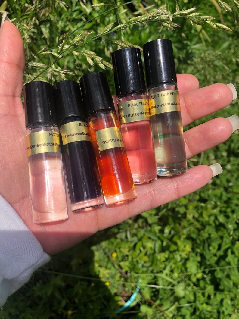 May include: Five small, clear glass roll-on bottles with black caps are held in a hand. The bottles contain various colored liquids, including pink, orange, and clear. Each bottle has a gold label with text, including "PUSS," "BLACK," "PINK SUGAR," and "BUTT NAKED."