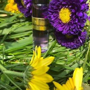 May include: A roll-on perfume bottle with a black cap and a gold label that reads "EAT IT RAW" and "butterMama". The bottle is surrounded by vibrant purple flowers with yellow centers and bright yellow sunflowers, all set against a backdrop of green grass.