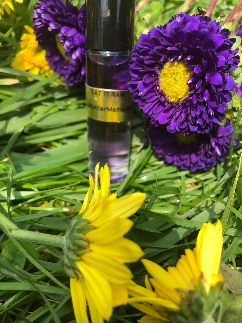 May include: A roll-on perfume bottle with a black cap and a gold label that reads "EAT IT RAW" and "butterMama". The bottle is surrounded by vibrant purple flowers with yellow centers and bright yellow sunflowers, all set against a backdrop of green grass.