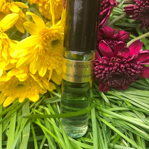 May include: A clear glass roll-on bottle with a black cap, labeled "Bob Marley" and "SheaButterMama", sits amidst yellow and burgundy flowers and green grass. The bottle contains a light green liquid.