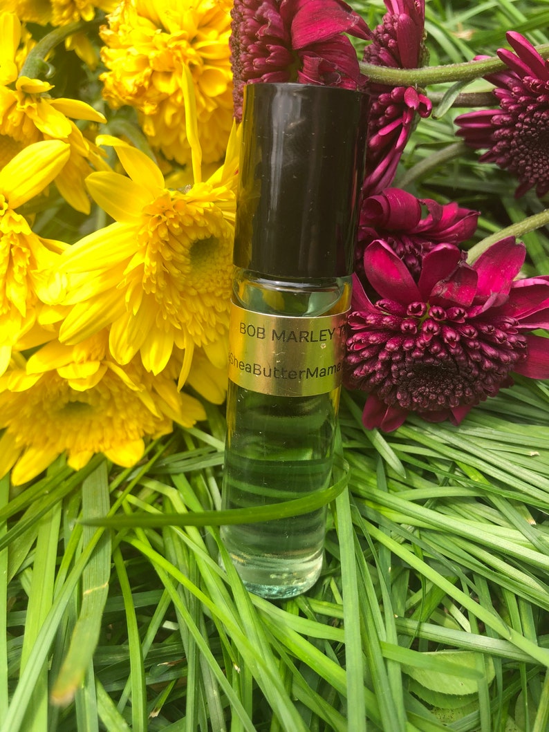May include: A clear glass roll-on bottle with a black cap, labeled "Bob Marley" and "SheaButterMama", sits amidst yellow and burgundy flowers and green grass. The bottle contains a light green liquid.