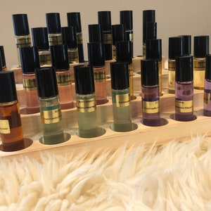 May include: A display of small glass roll-on bottles with black caps, filled with various colored liquids. The bottles are arranged in a wooden display rack. Each bottle has a gold label with text. The display sits on a fluffy, white surface.