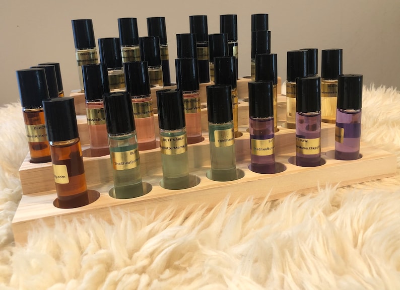 May include: A display of small glass roll-on bottles with black caps, filled with various colored liquids. The bottles are arranged in a wooden display rack. Each bottle has a gold label with text. The display sits on a fluffy, white surface.