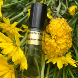 May include: A clear glass roll-on bottle with a black cap, labeled "Gucci Envy (M) Ty" and "SheaButterMama.E". The bottle is surrounded by bright yellow flowers and green foliage, creating a natural and vibrant setting.