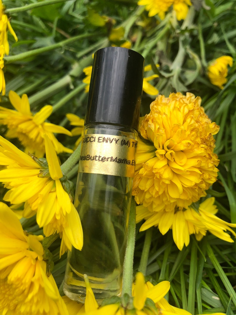 May include: A clear glass roll-on bottle with a black cap, labeled "Gucci Envy (M) Ty" and "SheaButterMama.E". The bottle is surrounded by bright yellow flowers and green foliage, creating a natural and vibrant setting.