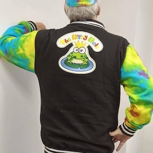 May include: Black varsity jacket with tie-dye sleeves and a matching tie-dye cap. The jacket features a cartoon frog graphic with the text "The Hippy Hole" on the back. The jacket has white stripes on the bottom and cuffs.