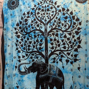May include: A blue and black tapestry featuring a detailed elephant design beneath a stylized tree. The elephant is adorned with intricate patterns, and the tree's leaves are heart-shaped. The background has a tie-dye effect.