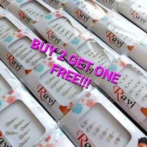 May include: A close-up of multiple packages of small, clear, adhesive rhinestones. The packages are white with pink accents and feature the text "Ravi Fancy Bindi" and "Buy 2 Get One Free!!!".