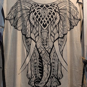 May include: A beige textile featuring a large, detailed black elephant design. The elephant's head and tusks are prominently displayed, with intricate patterns filling the ears and body. The edges are trimmed with a black border.