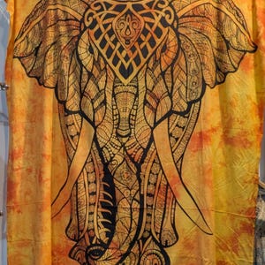 May include: A large, decorative tapestry featuring a detailed elephant design. The elephant is depicted in shades of yellow, orange, and black, with intricate patterns and a symmetrical composition. The background has a tie-dye effect, adding to the tapestry's artistic appeal.