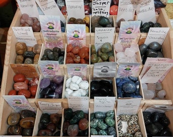 Puede incluir: Un expositor de madera lleno de diferentes piedras preciosas pulidas. Cada compartimento está etiquetado con el nombre de la piedra y sus propiedades. Las piedras están dispuestas en filas y columnas, con una variedad de colores y texturas.