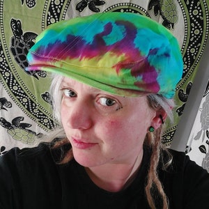 Peut inclure: Casquette plate tie-dye aux teintes de vert, bleu, violet et jaune. La casquette a une forme arrondie classique et un petit bord. En arrière-plan, une tapisserie verte avec des motifs de tortues.