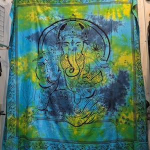 May include: A tie-dye tapestry featuring a detailed black illustration of the Hindu deity Ganesha. The tapestry has a vibrant color scheme of blue, yellow, and green. The border is adorned with a decorative floral pattern.
