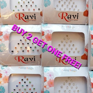 May include: Multiple packages of Ravi Fancy Bindi, each containing a collection of small, colorful, and clear rhinestones arranged in a triangular pattern. The packaging features floral designs and the text "Making Beauty". A promotional message reads "BUY 2 GET ONE FREE!"
