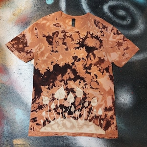 May include: A brown and tan tie-dye t-shirt with a white mushroom design. The short-sleeved top has a crew neck and a relaxed fit. The shirt is suitable for casual wear.