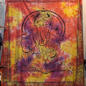 May include: A vibrant tapestry featuring a detailed illustration of the Hindu deity Ganesha. The artwork is rendered in black against a colorful backdrop of yellow, orange, and purple hues. The tapestry has a decorative border.
