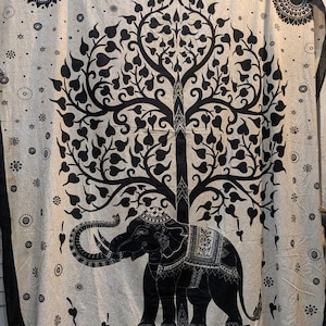May include: A beige and black tapestry featuring a detailed illustration of an elephant beneath a stylized tree. The elephant is adorned with decorative patterns. The design includes floral and geometric elements, with a black border.
