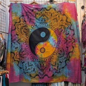 May include: A vibrant tapestry featuring a central Yin Yang symbol in black and tie-dye. The design is surrounded by a detailed mandala pattern in shades of pink, orange, yellow, and blue. The tapestry is displayed on a wall.