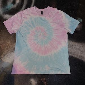 Könnte beinhalten: Kurzärmeliges T-Shirt mit einem Batikmuster in Hellblau und Rosa. Das Design zeigt ein Spiralmuster, das von der Mitte des Shirts ausgeht. Das Shirt hat einen Rundhalsausschnitt und ist aus einem weichen Material gefertigt.