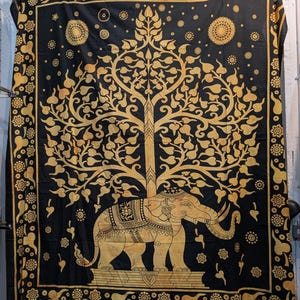 May include: Black and gold tapestry featuring an elephant beneath a tree. The design includes floral and sun motifs, with a decorative border. The elephant is detailed with ornate patterns. The tapestry is likely made of fabric.