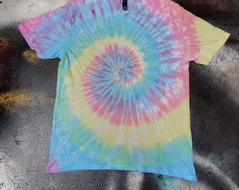 May include: A short-sleeved t-shirt featuring a spiral tie-dye pattern in pastel shades of pink, yellow, and light blue. The design radiates from the centre of the garment, covering the front with a blended, multi-coloured circular effect.