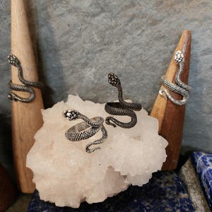 Op de afbeelding: Twee zilveren slangenringen op een witte kristallen steen. De ringen zijn gedetailleerd met schubben en hebben een ontwerp van een opgerolde slang.