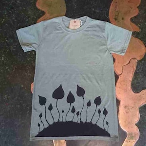 May include: A pale blue t-shirt with a black mushroom design. The graphic shows stylized mushrooms growing from a dark hill. The t-shirt has a round neck and short sleeves. A small tag is attached.