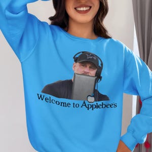 Puede incluir: Una sudadera azul brillante con un gráfico de un hombre que lleva una gorra de béisbol negra y auriculares. El gráfico está sosteniendo una tableta y el texto "Welcome to Applebee's" está impreso debajo del gráfico.