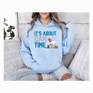 May include: Light blue hoodie with a white and blue graphic that says "It's About Dan Time" and a picture of a football player wearing a blue jersey with the word "Lions" on it.