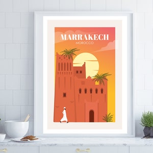 May include: Framed art print featuring a stylized illustration of Marrakech, Morocco. The artwork depicts a terracotta-colored building with palm trees, a setting sun, and the city's name in white text. A person in white clothing walks in the foreground.